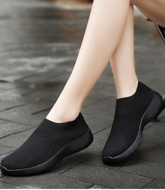 Black slip-on shoes worn by a person on a stone pavement.